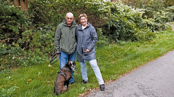 Zechenhäuser: Zwei Personen, ein Mann und eine Frau, nebeneinander, schauen in Kamera und vor ihnen ein großer Hund an der Leine, im Hintergrund Gebüsch