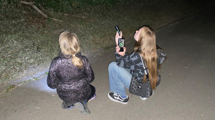 Zwei Mädchen hocken mit einem Ultraschalldetektor für Fledermäuse auf dem Boden.