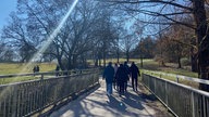 Von hinten fotografiert: Eine Gruppe Spaziergänger geht voraus, es sind Grünflächen und Bäume zu sehen. Die Sonne scheint, sodass sich ein Lichtstrahl im Bild widerspiegelt. 