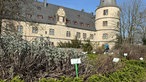 Die Gartenanlage der Wewelsburg. Im Vordergrund des Bildes ist ein Beet mit verschiedenen Pflanzen, darunter Salbei und Ysop, und entsprechend beschrifteten Schildern zu sehen, im Hintergrund sieht man die Wewelsburg. 