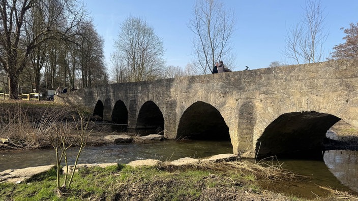 Der Fluss Alme fließt unter einer Brücke her. Man sieht zwei Personen auf der Brücke, weitere Spaziergänger gehen gerade auf die Brücke zu.