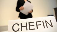 Frau im Hintergrund mit Dokumenten, vorne ein Schild "Chefin"