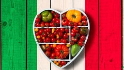 Verschiedene Tomaten und Paprika in einer herzförmigen Schachtel auf einem in den Farben der italienischen Flagge bemalten Holzuntergrund. 