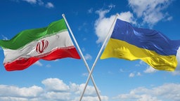 Symbolbild; zwei gekreuzte Flaggen der Ukraine und des Iran wehen im Wind vor einem bewölkten Himmel.