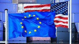 Fotomontage; EU- und US-Fahnen vor gestapelten Containern.