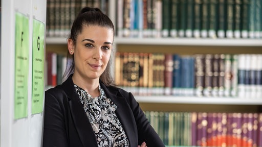 Dina El Omari steht mit verschränkten Armen in der Universitätsbibliothek und blickt freundlich in die Kamera