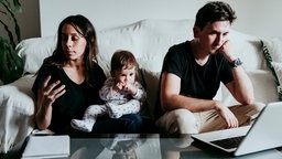 Mother using smart phone while father working on laptop with son sitting at home