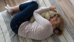 Eine Frau liegt in Fötusposition auf einem Bett und drückt ihr Gesicht in das Fell eines Kuscheltieres