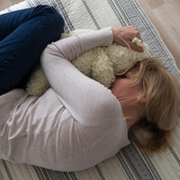 Eine Frau liegt in Fötusposition auf einem Bett und drückt ihr Gesicht in das Fell eines Kuscheltieres