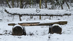 Eine Bank im Wald ist mit Schnee bedeckt. In den Schnee ist ein Herz gezeichnet.