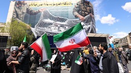 Teheran: Iraner mit Nationalflaggen schauen am 08.04.26 auf ein großes Banner mit der Aufschrift "Die Straße von Hormus bleibt geschlossen"