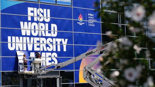 Mitarbeiter auf einer Hebebühne bekleben eine Fensterfront der Messe Essen mit Werbung für die World University Games. 