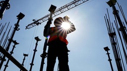 Ein Arbeiter auf einer Baustelle trinkt aus einer Flasche, im Hintergrund scheint die Sonne vom blauen Himmel.  
