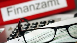  Auf einem Vordruck für die Steuererklärung liegt vor dem Aktenordner mit dem Aufdruck «Finanzamt» ein Stift und eine Brille.