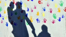 Ein Mann mit einem Kind auf dem Arm und einem an der Hand wirft einen Schatten auf eine mit bunten Handabdrücken bemalte Wand einer Kindertagesstätte