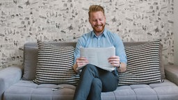 Mann sitzt auf einem Sofa und liest lächelnd die Zeitung. 