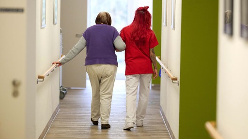 Symbolbild: Eine Pflegekraft führt eine Seniorin über einen Flur in einem Pflegeheim.