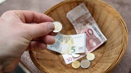 Symbolbild: Ein Mensch wirft eine Zwei-Euro-Münze in einen Korb mit Euro-Münzen und -Scheinen.
