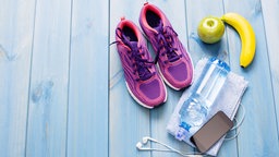 Sportschuhe, Handtuch, Wasserflasche Handy mit Kopfhörern und Obst auf einem blau gestrichenen Holzboden. Symbolbild