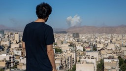 Ein Mann steht auf einem Aussichtspunkt und blickt auf das Stadtpanorama von Teheran, im Hintergrund ist Rauch zu sehen. 