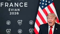 Symbolbild: US-Präsident Trump vor dem Logo des G7-Gipfels 2026 in Evian und einer US-Flagge.