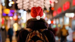 Symbolbild: Rückansicht eines Menschen mit Weihnachtsmütze in einer weihnachtlich beleuchteten Einkaufstraße.