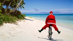 Symbolbild: Ein als Weihnachtsmann verkleideter Mensch fährt an einem Palenstrand Fahrrad.