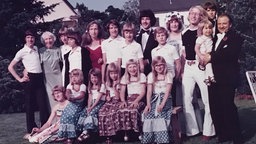 Eine Großfamilie in den 70- oder 80-iger Jahren posiert für ein Gruppenfoto im Garten. 