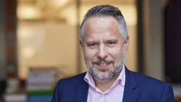 Porträtaufnahme von Andreas Peichl: Er hat braune Augen, kurzes, graues, nach hinten gekämmtes Haar, einen grauen Vollbart und trägt ein blaues Sakko mit einem rosafarbenen Hemd.