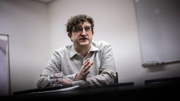 Das Foto von Andreas Stahl zeigt ihn sitzend an einem Tisch: mit kurzem lockigem Haar, dunkler Brille und hellem Hemd.