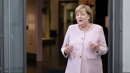 Angela Merkel steht vor einer offen Tür und gestikuliert, während sie in die Kamera blickt.