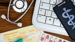 Symbolfoto: Ein Stethoskop, eine Computertastatur, ein Smartphone und eine Überweisung liegen auf einem Tisch.
