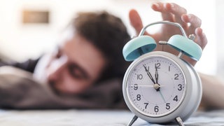 Im Vordergrund ein türkiser Wecker, der von einem müden Mann in einem Bett ausgeschaltet wird.
