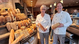 Zwei Bäcker unterschiedlichen Alters, Günther und Sebastian van Densen aus Tönisvorst, stehen in einer Bäckerei neben einer Brotauslage und halten jeweils ein Brot in die Kamera.