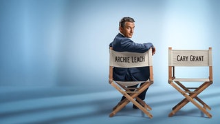 Jason Issacs sitzt in seiner Rolle als Archie Leach auf einem Regisseurstuhl, der mit der Rückseite zur Kamera steht. Er ist mit einem Arm auf die Lehne gestützt,blickt nach hinten gedreht in die Kamera. Daneben steht noch ein Cary Grant-Regisseurstuhl 