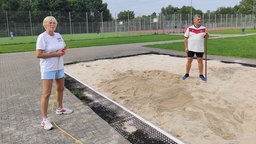 Die Prüfer Rita und Dieter Putzke stehen auf einem Sportplatz an bzw. in einer Sandgrube