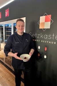 Sternekoch Egor Hopp trägt kurzes, nach hinten gekämmtes Haar, eine schwarze Küchenuniform und ein breites Lächeln im Gesicht.