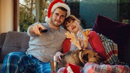 Ein junges Paar sitzt in der Weihnachtszeit mitsamt Hund gemütlich auf dem Sofa und schaut fern.