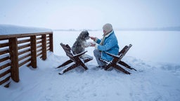Frau und ihr Hund sitzen auf Holzstühlen in der Nähe des schneebedeckten, zugefrorenen Sees Pudasjärvi, Nordösterbotten, in Finnland.