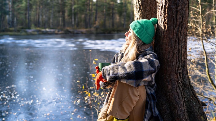 Eine Frau lehnt an einem Baum an einem See im Winter und genießt die Ruhe. 