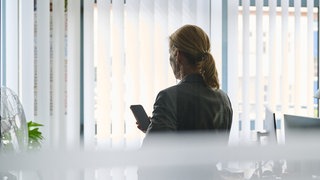 Eine Frau in Business-Kleidung steht in einem Büro und führt ein Gespräch über ihr Smartphone.