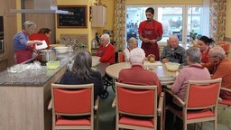 Bewohner und Mitarbeiter stehen und sitzen im Haus Rheinaue und bereiten gemeinsam Essen zu