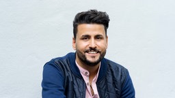 Die Porträtaufnahme von İbrahim Arslan zeigt ihn freundlich lächelnd mit gestyltem, dunklem Haar, Vollbart, einer blauen Jacke und einem rosafarbenen Hemd.