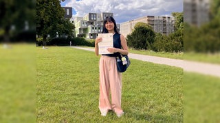 Eine junge Frau steht auf einer Wiese und hält ihre Wahlbenachrichtigung zur Integrationsratswahl in der Hand.