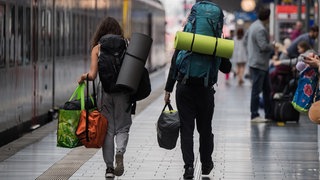 Zwei Menschen mit schwerem Outdoor-Reisegepäck auf den Schultern gehen über den Bahnsteig zu einem Zug.