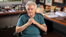 Das Foto zeigt John Irving, der auf einem Bürostuhl in seinem Arbeitszimmer sitzt und die Hände auf Brusthöhe faltet.