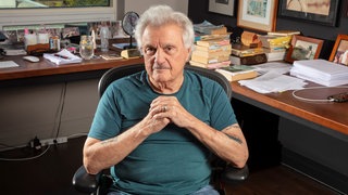 Das Foto zeigt John Irving, der auf einem Bürostuhl in seinem Arbeitszimmer sitzt und die Hände auf Brusthöhe faltet.