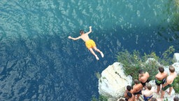 Junger Mann springt von einem hohen Felsen mit Zuschauern ins Wasser