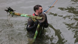Ein Mann in einem Neoprenanzug, an einem Seil gesichert, birgt einen mit Matsch bedeckten E-Scooter aus dem Rhein.