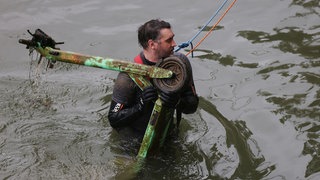Ein Mann in einem Neoprenanzug, an einem Seil gesichert, birgt einen mit Matsch bedeckten E-Scooter aus dem Rhein.